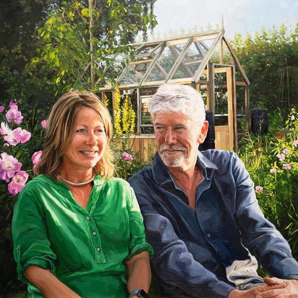 Michael and Sally A commissioned oil portrait painting of a smiling couple seated outdoors among flowers, with a greenhouse in the background in the uk.